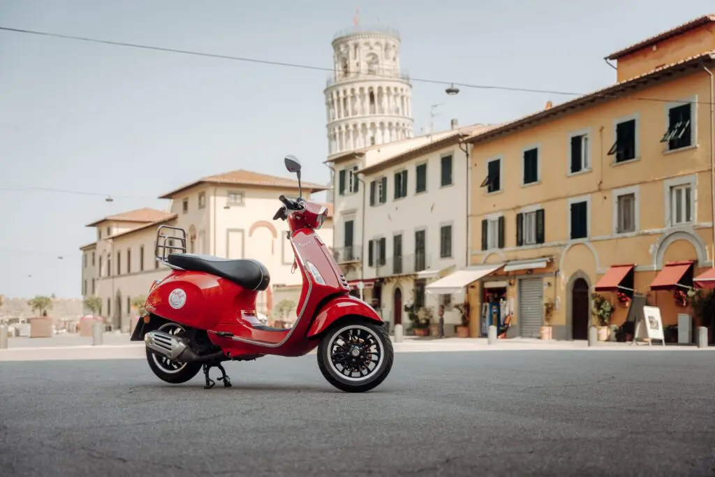 red Vespa scooter parked in a piazza with the Leaning Tower of Pisa