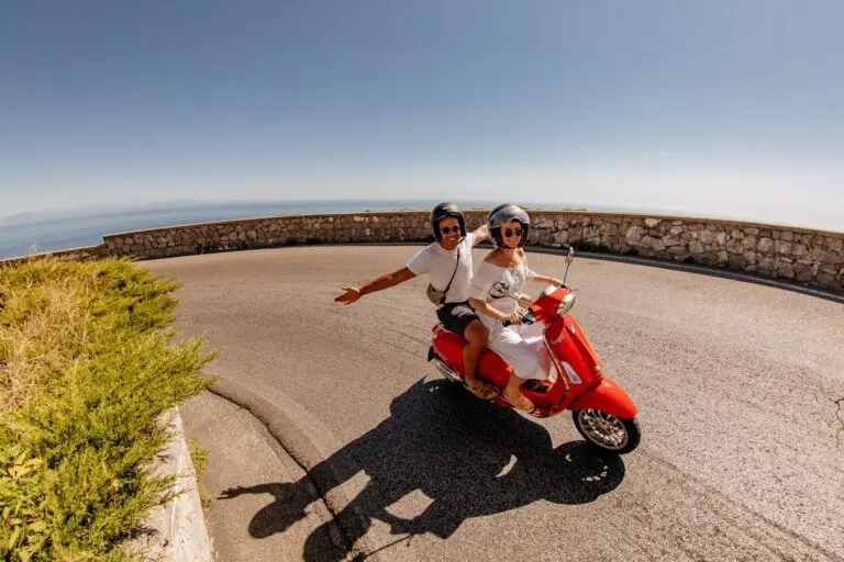 couple riding red Vespa scooter on coastal road
