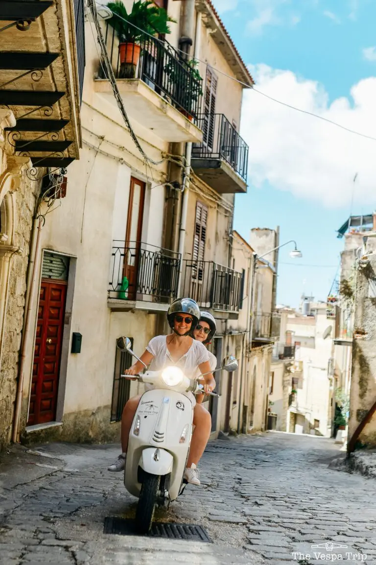 two women riding a white Vespa scooter on a cobbled street in Sicily