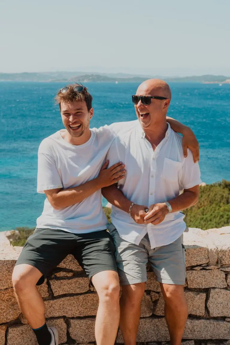 two men laughing on a stone wall overlooking the sea in Sardinia